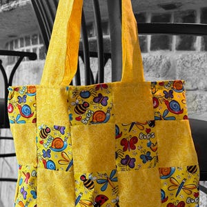 May include: A yellow tote bag with a patchwork design featuring colorful insects and snails. The bag has yellow straps and is made of fabric. The background is a black and white image of a chair and wall.