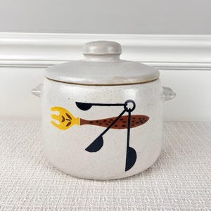 May include: A white ceramic crock with a lid, featuring a brown, yellow, and black design of a fork, spoon, and ladle. The crock has a speckled texture.