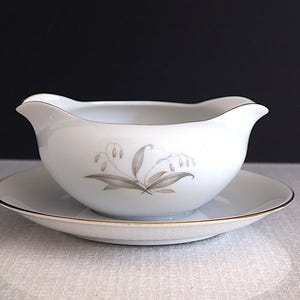 May include: A white porcelain gravy boat with a floral design and a matching saucer. The gravy boat has a wide, rounded base and a flared lip. The saucer is round and has a slightly raised edge.