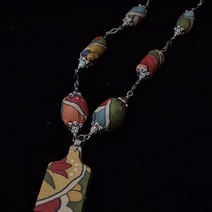 Peut inclure: Un collier en tissu coloré avec un pendentif rectangulaire et des perles décoratives. Le pendentif et les perles présentent des motifs rouges, jaunes, verts et bleus. Le collier est sur une chaîne argentée.