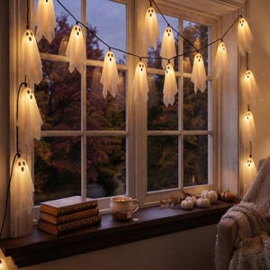 May include: A string of white ghost-shaped LED lights hanging across a window frame. Each ghost features a simple black face with two eyes and a mouth. The lights emit a warm glow, illuminating the translucent fabric draped over each individual bulb.