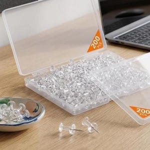 May include: A clear plastic storage container filled with numerous transparent push pins, featuring a hinged lid and an orange triangular label marked with 200 PCS. Several additional clear push pins are scattered on the surface nearby.