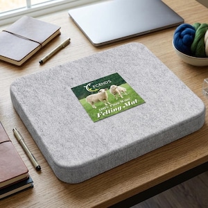 May include: A gray felt felting mat with a label that reads "100% Pure Wool Felting Mat" and features two sheep. The mat is on a wooden desk with a laptop, notebooks, pens, and a small bowl of wool roving.