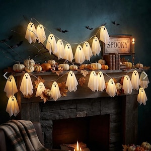 May include: A string of white ghost-shaped LED lights draped across a fireplace mantel decorated with small pumpkins, autumn leaves, and a sign that reads SPOOKY. The ghost lights feature simple black eyes and mouths, emitting a warm glow.