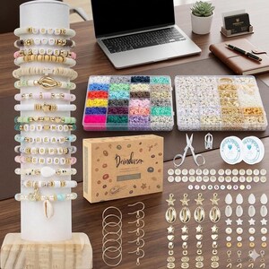 Puede incluir: Un kit para hacer joyas que incluye un expositor cil&iacute;ndrico blanco con varias pulseras de cuentas, dos organizadores de pl&aacute;stico transparente con cuentas de arcilla y met&aacute;licas, una caja de cart&oacute;n con la etiqueta Deinduser, tijeras y varios dijes.
