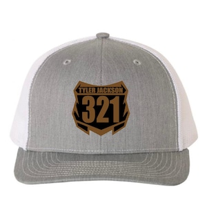 May include: A gray and white trucker hat with a brown and black shield-shaped emblem. The emblem features the text "TYLER JACKSON" above the number "321". The hat has a curved brim and a mesh back.