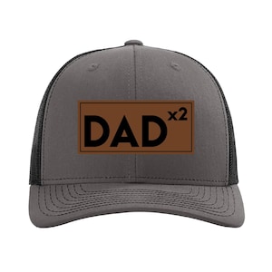 May include: A gray and black trucker hat with a brown leather patch that says "DAD x2" in black text.