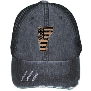 May include: A black distressed denim trucker hat with a black mesh back. The hat features a distressed American flag design in the shape of the state of Vermont.