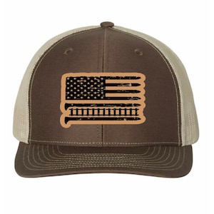 May include: A brown and tan trucker hat featuring a distressed American flag design. The flag graphic is black and tan, with a star field and stripes. The hat has a mesh back and a curved brim.