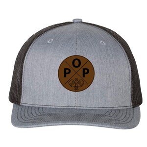 May include: Gray and black trucker hat with a brown circular patch. The patch features the letters "O", "P", and "P" in a cross design, along with a pickleball and paddles. The hat has a curved brim and a mesh back.
