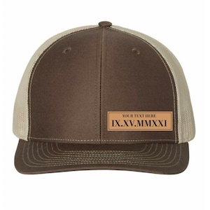 May include: A brown trucker hat with a tan mesh back. The front panel is brown with a rectangular patch that reads "YOUR TEXT HERE" above the Roman numerals "IX.XV.MMXXI". The hat has a curved brim.