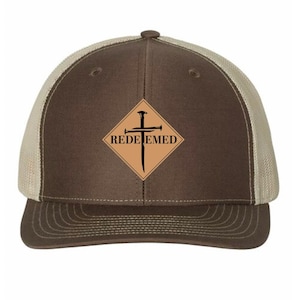 May include: A brown and tan trucker hat with a mesh back. The front features a diamond-shaped patch with the word "REDEEMED" and a cross and nail design in black. The hat has a curved brim and a button on top.