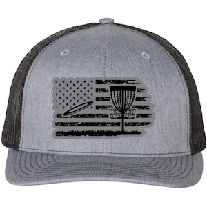 May include: A gray and black trucker hat with a distressed American flag design featuring a disc golf basket and disc. The flag is black and white with a gray background.