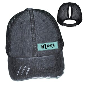 May include: Distressed black trucker hat with a mesh back and a curved brim. The hat features a teal rectangular patch with the name "Mimi" in a cursive font. The hat has a ponytail opening at the back.