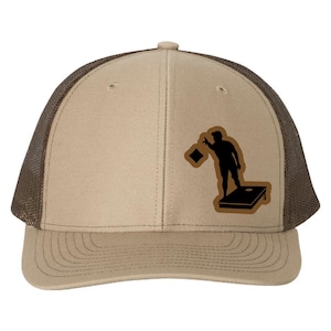 May include: Tan and brown baseball cap with a mesh back. The cap features a black silhouette of a person playing cornhole, throwing a bean bag. The design is on a brown patch.