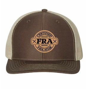 May include: A brown and tan trucker hat with a curved brim. The hat features a circular patch with the words "CERTIFIED FRAX VIOLATOR" in black and tan. The patch includes crossed hammers.