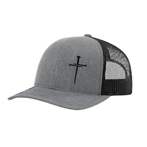May include: Gray trucker hat with a black mesh back and a curved brim. The hat features a black cross design on the front. The brim has dark blue stitching.