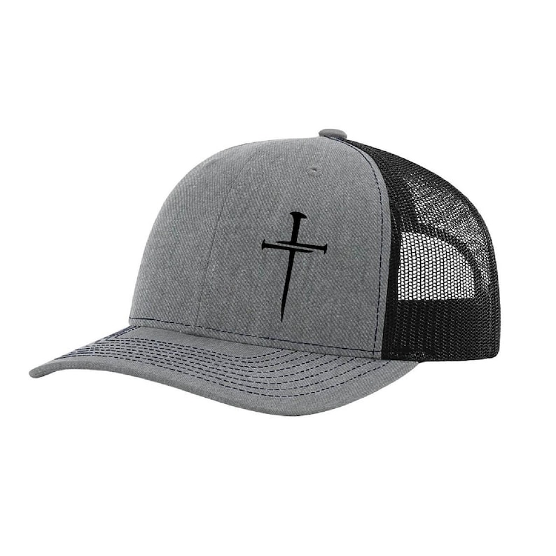 May include: Gray trucker hat with a black mesh back and a curved brim. The hat features a black cross design on the front. The brim has dark blue stitching.