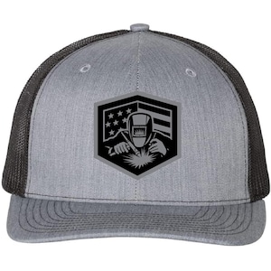 May include: A gray and black trucker hat with a black and white graphic of a welder wearing a welding mask and holding a welding torch. The graphic is set against an American flag background.