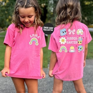 A young girl wearing a pink t-shirt with various colorful and whimsical illustrations, including a rainbow, flowers, and a character.