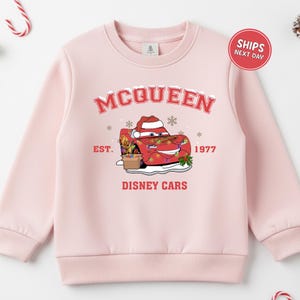 A pink sweatshirt with a graphic design featuring a character from the Disney movie Cars, along with text and a red circular sticker indicating that the sweatshirt is for shipping next day.