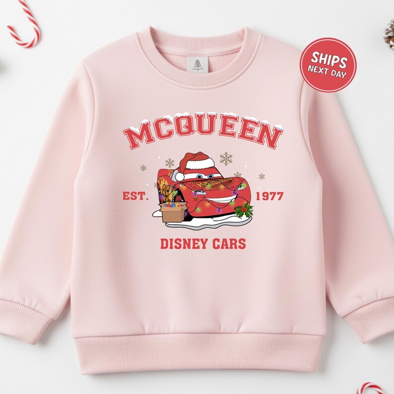 A pink sweatshirt with a graphic design featuring a character from the Disney movie Cars, along with text and a red circular sticker indicating that the sweatshirt is for shipping next day.