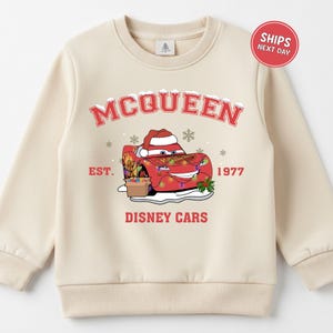 A beige sweater with a graphic design featuring a red car character from the Disney movie Cars, along with snowflakes and text.