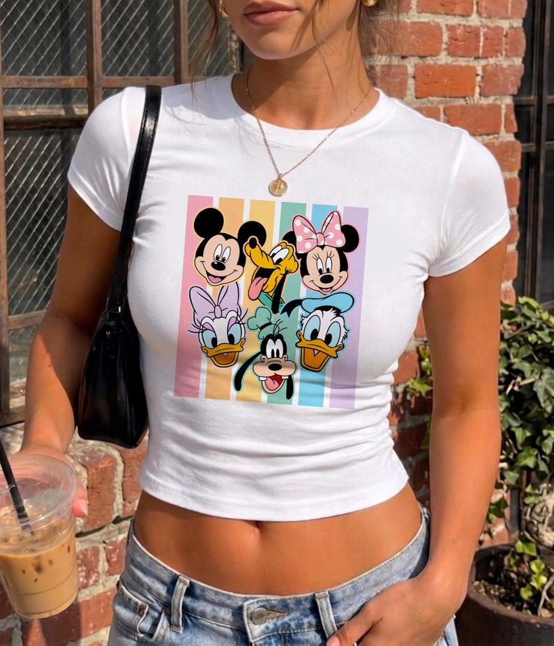 A woman wearing a white t-shirt with a graphic of Disney characters Mickey Mouse, Minnie Mouse, Donald Duck, Goofy, and Pluto. She is holding a drink in her hand.