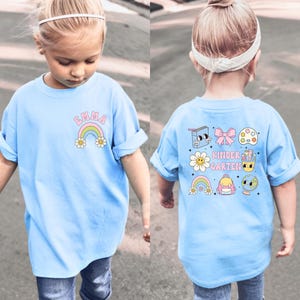 A young girl wearing a blue t-shirt with a rainbow and cartoon characters on the back, walking on a street.