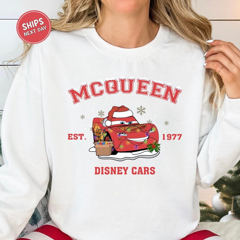 A white sweatshirt with a graphic design featuring a red car and snowflakes, along with the text "McQueen" and "Disney Cars" printed on it.