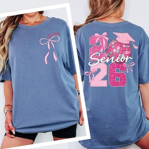 A woman wearing a blue t-shirt with a pink ribbon and the text "Emma 26" printed on it.