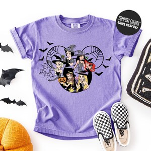 A purple t-shirt with a Halloween-themed graphic design featuring characters from Disney's "Snow White and the Seven Dwarfs" and bats. The t-shirt is displayed on a white background, accompanied by a pair of black and white checkered shoes, a black bat decoration, and a yellow hat.