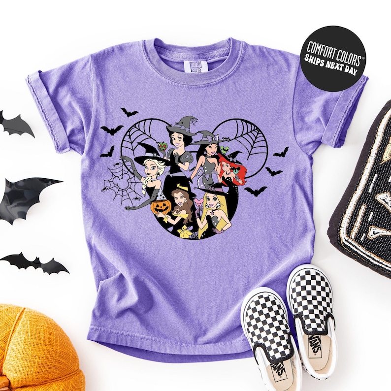 A purple t-shirt with a Halloween-themed graphic design featuring characters from Disney's "Snow White and the Seven Dwarfs" and bats. The t-shirt is displayed on a white background, accompanied by a pair of black and white checkered shoes, a black bat decoration, and a yellow hat.