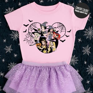 A pink t-shirt with a cartoon design of a group of girls dressed in Halloween costumes, including a witch, a pumpkin, and other spooky elements, along with a purple tutu skirt.