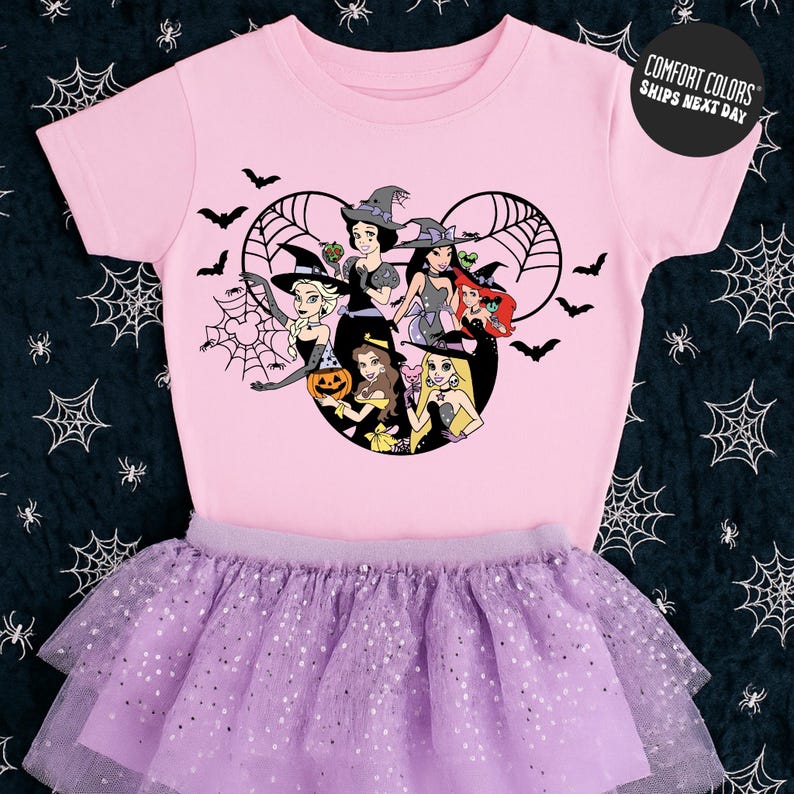 A pink t-shirt with a cartoon design of a group of girls dressed in Halloween costumes, including a witch, a pumpkin, and other spooky elements, along with a purple tutu skirt.