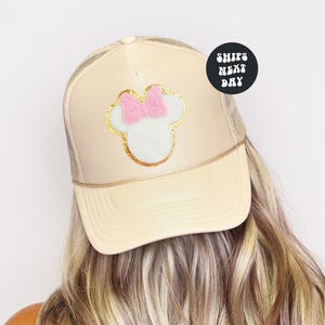 May include: Cream-colored trucker hat featuring a Minnie Mouse design. The design includes a pink bow, white face, and gold glitter accents. The hat has a mesh back and a curved brim. The text "SHIPS NEXT DAY" is on a black circle.