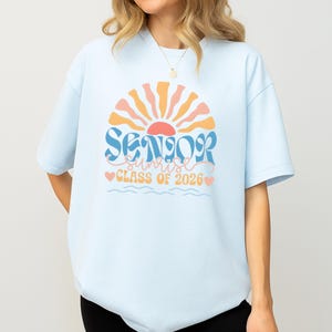 A woman wearing a light blue t-shirt with a graphic design that says "SENIOR SUNSET CLASS OF 2026".