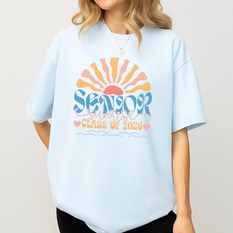 A woman wearing a light blue t-shirt with a graphic design that says "SENIOR SUNSET CLASS OF 2026".