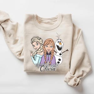 A beige sweater with a graphic design featuring two cartoon characters, a blonde girl and a snowman, along with the name "Olivia" written below.