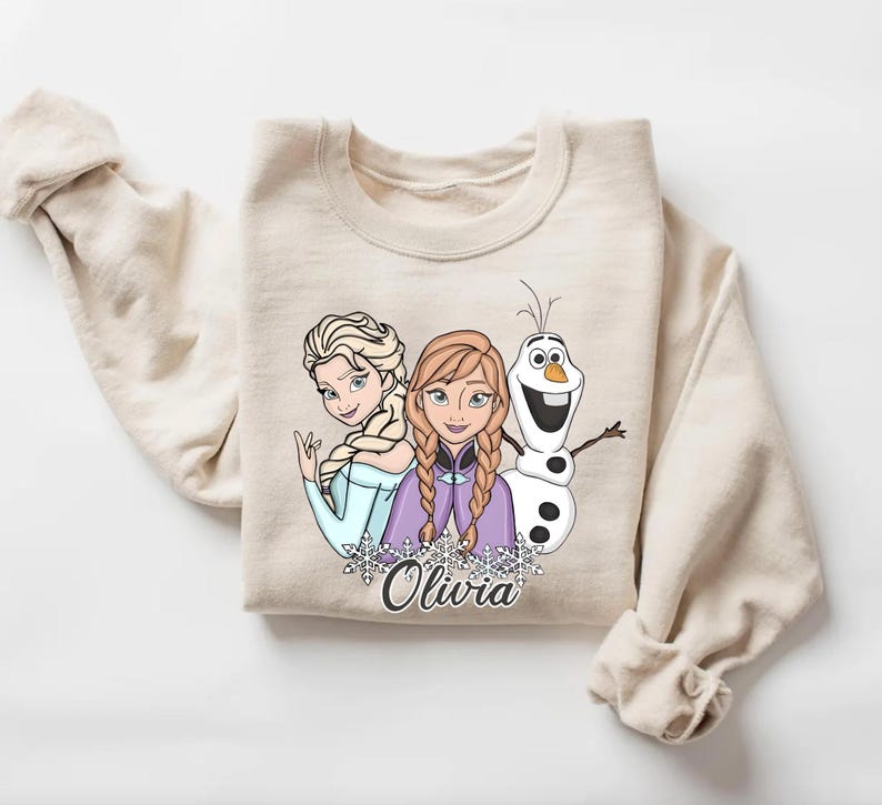 A beige sweater with a graphic design featuring two cartoon characters, a blonde girl and a snowman, along with the name "Olivia" written below.
