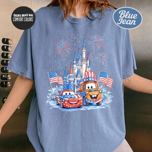 A person wearing a blue t-shirt with a graphic design featuring two cartoon cars, one orange and one blue, along with a castle and fireworks in the background.