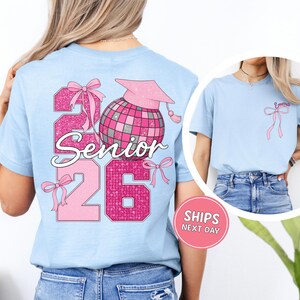 A woman wearing a light blue t-shirt with a graphic design on the back, featuring a pink and purple color scheme and the text "26 Senior" along with a pink and purple disco ball and ribbon.