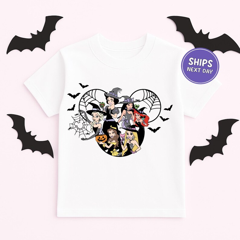 A white t-shirt with a graphic design featuring cartoon characters, including a witch, a pumpkin, and other Halloween-themed figures, surrounded by bats.
