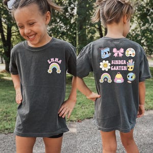 A young girl wearing a gray t-shirt with colorful cartoon characters and text on the back.