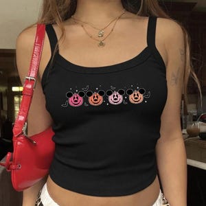 May include: A black ribbed tank top featuring a graphic print of four pumpkin characters with mouse ears in shades of pink and orange, accented by small bats and stars. A bright red shoulder bag with a silver buckle is visible on the side.