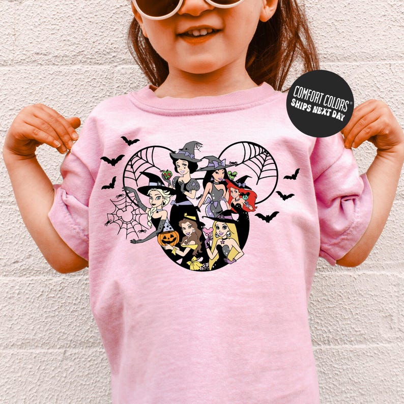 A young girl wearing a pink t-shirt with a graphic design featuring cartoon characters, including a witch, a pumpkin, and other Halloween-themed figures.