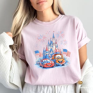 A woman wearing a pink t-shirt with a graphic of two cartoon cars, one red and one orange, holding American flags, and a castle in the background.