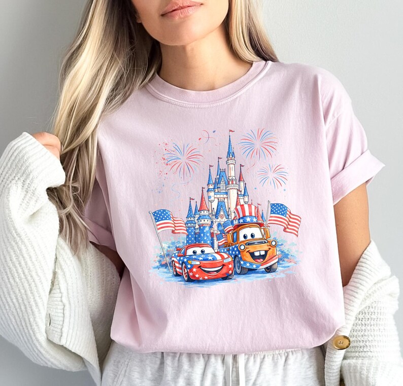 A woman wearing a pink t-shirt with a graphic of two cartoon cars, one red and one orange, holding American flags, and a castle in the background.