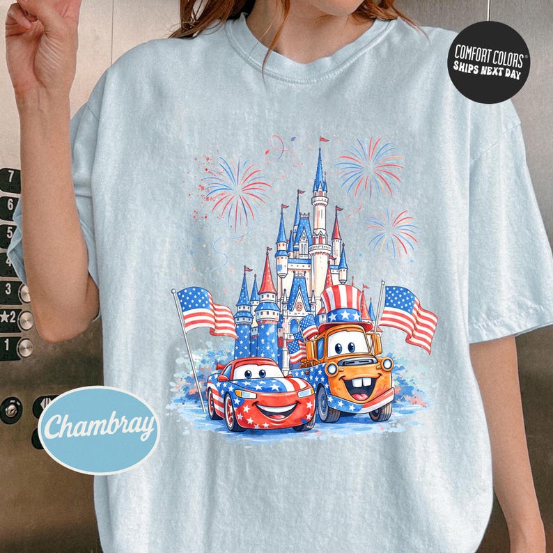 A person wearing a white t-shirt with a graphic design featuring a castle, two American flag-themed cars, and fireworks in the background.