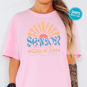 A woman wearing a pink t-shirt with a graphic design that says "SENIOR CLASS OF 2026" and features a sun-like design.